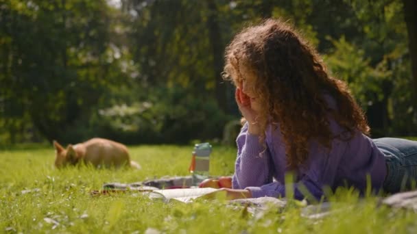 白种人快乐的女孩饲养员宠物主人在草地上看书夏天周末带着顽皮的小狗在公园里笑着年轻的女学生在外面看书放松休息 — 图库视频影像