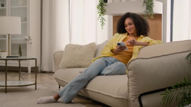 ハッピー笑顔のアフリカ系アメリカ人女性エスニックガール 快適なソファー居心地の良いソファーでチャットオンライン使用 リビングルームの快適さで携帯電話ガジェットスマートフォンで中毒ソーシャルメディアをスクロールするモバイルアプリ — ストック動画