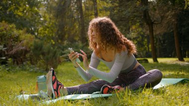 Kafkasyalı cihaz bağımlısı sosyal medya cep telefonu bağımlısı kadın sporcu kız spor antrenmanı vücut esnetme egzersizi yoga minderi açık havada sarkan akıllı telefon etkileyicisi