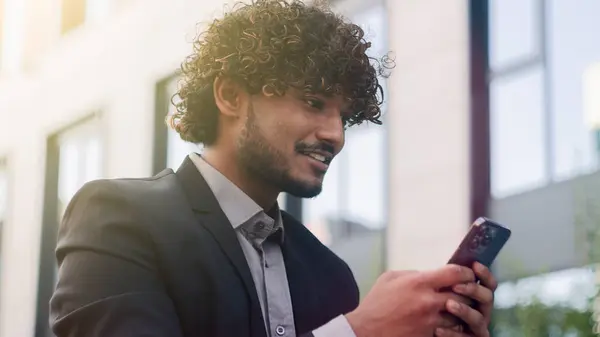 Indian employer Arabian entrepreneur happy amazed cheerful positive businessman using smartphone outdoors in city business man chatting with mobile phone online reading good news corporate success