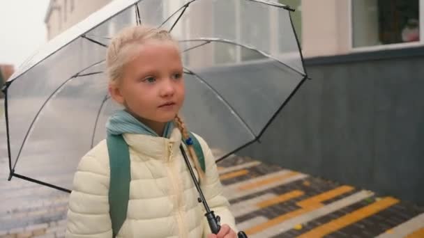 小さなヨーロッパのかわいい少女の子供は雨の雨の天気の外の街路を歩いています カフカシアの子供の生徒は 屋外で雨の日に小学校に行く周りを見回しています — ストック動画