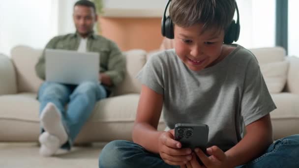 小男孩在手机上玩电子游戏在地板上快乐的多种族家庭在家里使用各种各样的设备妈妈爸爸在网上玩笔记本电脑背景小孩玩智能手机 — 图库视频影像