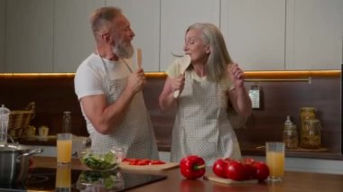 Orta yaşlı emekli çift, mutlu beyaz aile babası yaşlı eş mutfakta birlikte şarkı söylüyor komik sağlıklı sebzeler pişiriyor olgun kadın ve erkek mutfak kaşıklarında şarkı söylüyor.