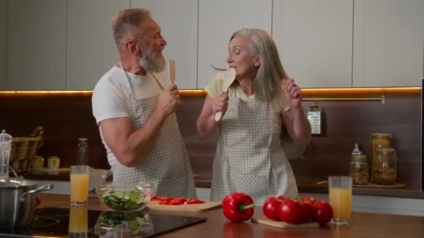 中年引退したカップルコーカサスハッピーファミリー夫妻はキッチンで楽しい歌を歌って 面白い料理新鮮な健康な野菜成熟した女性と男は台所用品のスプーンで歌います — ストック動画