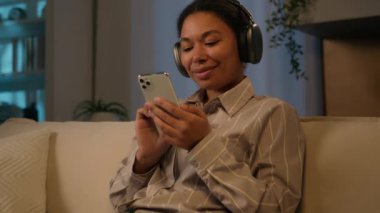 Rahatlamış gülümseyen Afrikalı kadın, kulaklıklı Etnik Amerikalı kız, evde rahat bir gece evde rahat müzik, podcast dijital uygulama servisi, cep telefonu medyası.