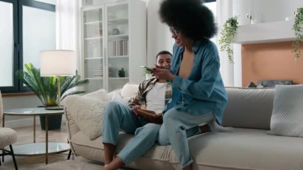非洲裔美国男人在家里的沙发上看书快乐而又兴奋的女人跑进客厅 在手机上展示自己的出价 分享好消息夫妇在智能手机上的成功 — 图库视频影像