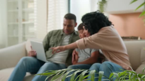 自宅のソファーでラップトップコンピュータを使用して幸せな多種多様な家族の両親の子供は興奮するアフリカ系アメリカ人のお父さんコーカサス州養子の息子は自宅で勝利の成功を祝います — ストック動画