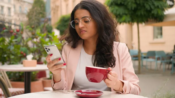Unhappy young Indian Arabian ethnic woman female girl drinking coffee feel annoyed negative message notification mobile phone outside city street cafe. Overworking problem online gadget error trouble