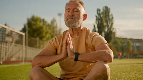 Old European man closed eyes meditating on grass in city relaxation rest healthy body mind elderly sportsman outdoors pray yoga zen balance peace practice energy pose lotus outdoors well-being sport