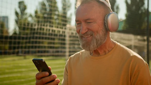 Caucasian happy old man walking stadium football field with headphones listening music smiling texting using phone smartphone cellphone social media outside in city pensioner sport messages enjoyment