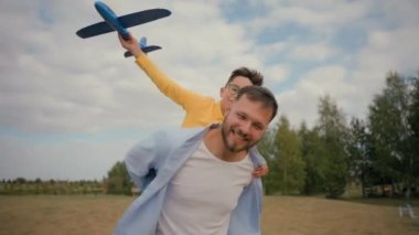 Mutlu babalık ailesi, gülümseyen beyaz erkek baba, neşeli oğul, sırtına binen baba, oyuncak uçak oynayan çocuk pilot, kır parkının dışında iyi uçuşlar.