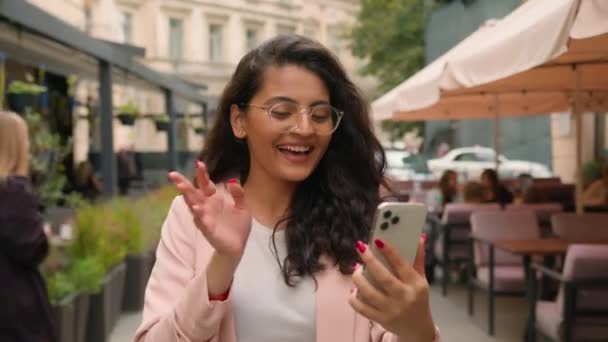 ハッピー笑顔のインドのアラビアの民族女性女性のビジネスマン観光客は 市街地のカフェの外で携帯電話のカメラを振って挨拶するビデオ通話をする ビジネスオンライン接続アプリ — ストック動画