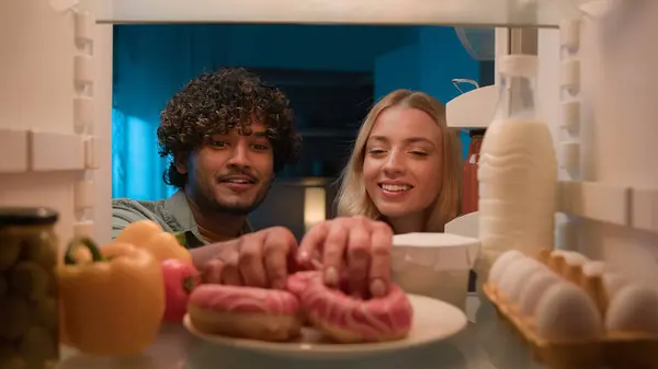 Point of view POV from inside refrigerator multiracial multiethnic couple eat tasty donuts happy enjoy junk food at home kitchen together eating Indian man and Caucasian woman at night open fridge