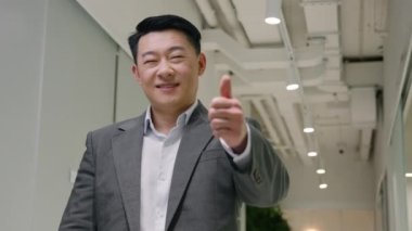 Memnun Asyalı, mutlu, orta yaşlı, Koreli, Çinli, etnik, erkek, girişimci, yönetici, ofis koridorlarında iş başarısı misali baş parmağını gösteriyor.