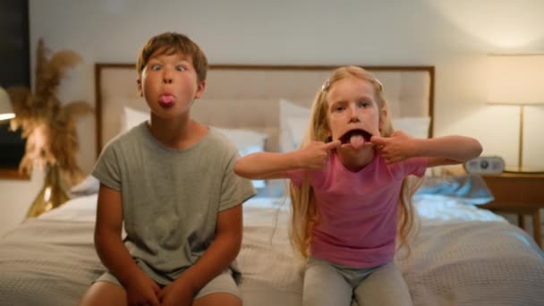 おかしな幸せなコーカサス人の子供の少年少女の兄弟姉妹の息子の娘の友人は 寝室で舌の冗談を示す顔を作るために一緒に遊んで笑う家でカメラを引き裂きました — ストック動画