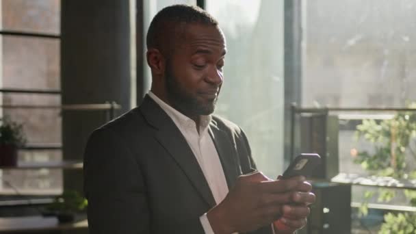 在阳光灿烂的办公室里微笑着成熟的商人用手机体贴的非洲裔美国人职业经理人在线聊天手机数字应用思考搜索业务解决方案微笑 — 图库视频影像