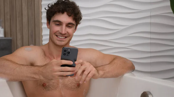 Relaxed man in bath relaxing washing in bathtub foamy hot water talking to mobile phone web camera video call distant chat record vlog blog male influencer talk to smartphone social media say bye
