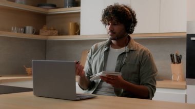 Uzaktan e-öğrenim ev eğitimi alan, kulaklık takmış, bilgisayar sınıfı dizüstü bilgisayarı izleyen, Hintli erkek öğrenci internet öğretmeni, not yazıyor, SAT sınavı kursuna hazırlanıyor.