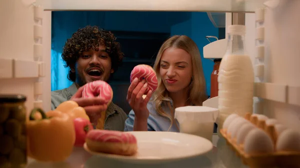 Point of view POV from inside refrigerator multiracial couple diverse Indian guy man and Caucasian girl woman at night kitchen open fridge eating donut unhealthy nutrition diet failure obesity food