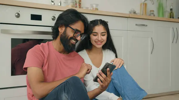 Arabian Indian interracial couple woman man sit on warm floor in kitchen watching funny video content in social media ordering online comfort delivery services hold mobile phone buy goods on internet