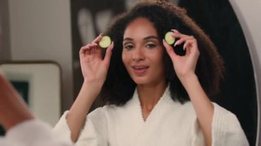 Mutlu gülümseyen sağlıklı Afro-Amerikan kadın banyodaki spa salonunda elinde iki halka salatalık dilimi ile kameraya bakan ayna yansıması gözlere bakıyor.