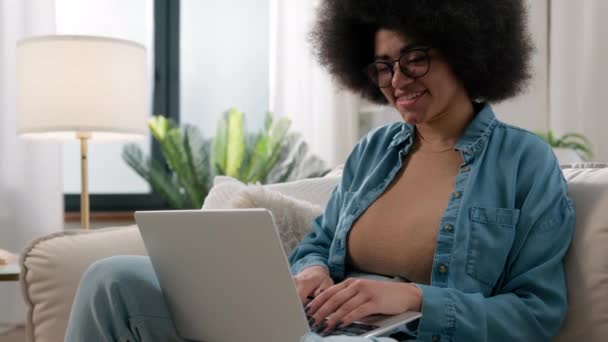 ソーシャルメディアのテキストで友人とチャットする自宅でラップトップを持つアフリカ系アメリカ人女性エスニックガール コンピュータで笑顔のニュース オンラインショッピングアプリを購入ソファでPcを使用して幸せ — ストック動画