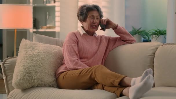 舒舒服服地在舒适的沙发上笑着的老妇人用智能手机笑着谈论有趣的新闻无忧无虑地在家里舒服的沙发上老年妇女退休后说手机 — 图库视频影像
