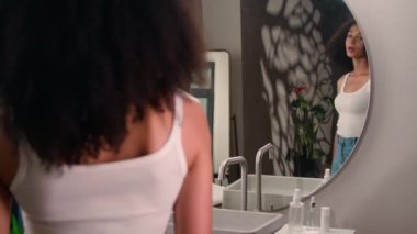 Afro-Amerikalı bir kadın banyoya gelir aynanın yansımasına bakar güzel etnik kız kız bukleleri düzeltir kıvırcık saçlar yapar saç stili şampuanı yapar, rutin güzellik şampuanı gider.
