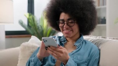 Mutlu Afrikalı Amerikalı kız elinde cep telefonu cihazıyla evinde gülümseyen genç bir kadın cep telefonu kullanıyor kadın nüfuzlu blogcu sosyal medya bağımlısı internetten satın alıyor.