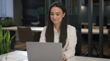 Ofis masasında dizüstü bilgisayarla çalışan, motive olmuş, beyaz, gülümseyen bir iş kadını mutlu bir iş kadını bilgisayarla çalışıyor ve işini iyi yaptığını düşünüyor.