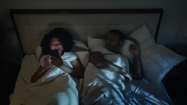 アフリカ系アメリカ人のガジェット中毒ソーシャルメディアの携帯電話中毒女性 ベッドの夜にスマートフォンを使用して笑う怒った男 夫は枕の騒々しい笑いで妻のカバーヘッドをプッシュ — ストック動画