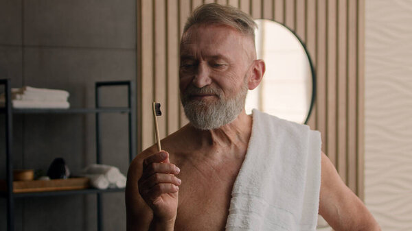 Old Caucasian retired male senior man with bath towel on shoulder showing holding bamboo tooth brush recommend brushing teeth oral care clean health mouth hygiene dental morning habit routine bathroom
