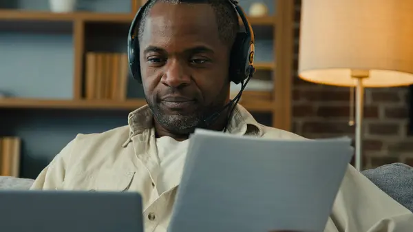 African american man teacher businessman freelancer in headset checking documents entering data in laptop remote working at home using computer application paying bills processing online information