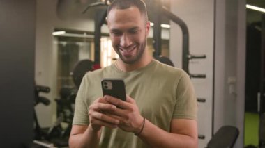 Amerikalı Latin erkek sporcu formda sporcu fitness eğitmeni vücut geliştirme sporcusu erkek cep telefonu kullanarak akıllı telefon uygulamasında sosyal medyada mutlu bir gülümseme haberi veriyor.