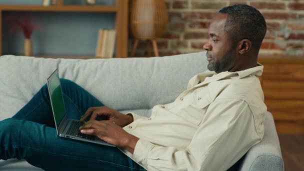アフリカ系アメリカ人男性ビジネスマンフリーランサー ラップトップブラウジングメディアネットワークでタイプするリビングルームのソファーに横たわるソファでリラックスするコンピュータアプリを使用してオンラインで電子メールメッセージを送信します — ストック動画