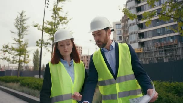 承包商多样性工程师两个人同事男建筑商身穿安全制服戴防护帽头戴安全帽的建筑师在谈论城市的蓝图建筑平面图 — 图库视频影像