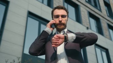 Meşgul yetişkin iş adamı Kafkasyalı cep telefonundan konuşan akıllı telefon görüşmesi ofis dışından erkek girişimci CEO bilek saatine bakıyor şehir caddesinde müşteri işleri toplantısı bekliyor.
