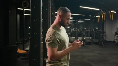 Amerikalı Latin kökenli bir adam akıllı telefonlara bakıyor sporcu formundaki sporcu spor eğitmeni spor salonundaki vücut geliştirme sporcusu cep telefonuyla kameraya bakıp mutlu bir şekilde gülümsüyor.