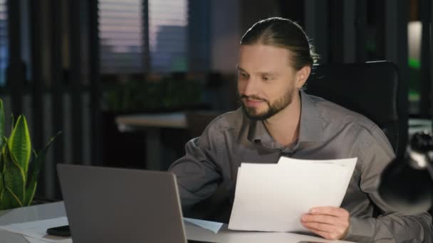 夜のオフィスの仕事のペーパーでコーカサス人のビジネスマンは文書を確認しますノートパソコンのペーパーワークを入力するデータ 満足したビジネスマンの起業家ブックキーパー 企業の財務をチェック — ストック動画