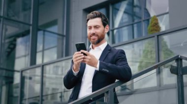 Başarılı gülümseyen Kafkasyalı iş adamı lider ofis binasının yakınındaki telefonu kullanarak sosyal medyada geziniyor e-postaları kontrol ediyor akıllı telefondan mesajlar gönderiyor.