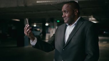 Öfkeli hoşnutsuz Afro-Amerikan iş adamı, karanlık yeraltı otoparkında cep telefonuyla video görüşmesi yapan işadamı akıllı telefon webcam kullanarak çatışma stresi sorununu çözüyor.