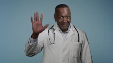 Afro-Amerikalı dost canlısı, gülümseyen erkek doktor, doktor doktor doktor, tıbbi uzman, mavi arka plan stüdyosunda kameraya bakıyor, merhaba reklam kliniği, sağlık hizmeti konseptini selamlıyor.