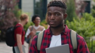 Mutlu Afrikalı Amerikalı öğrenci erkek öğrenci açık hava üniversitesi kampüsünde kameraya gülümseyen erkek öğrenci eğitim çalışmalarının keyfini çıkarın arka planda konuşan Z kuşağı öğrencileri