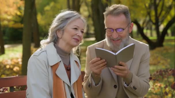 退休的白种人夫妇白发苍苍的祖父母在秋天的公园里看书养恤金闲暇时间快乐的70多岁的爱丈夫和妻子一起坐在长椅上休息户外退休 — 图库视频影像