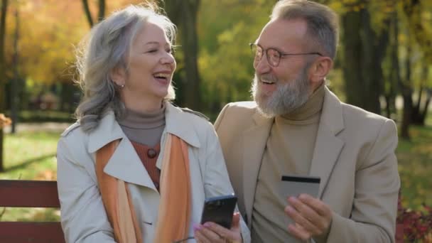 快乐的白种人已婚夫妇成熟的老妇人祖父母使用智能手机信用卡银行应用软件网上购物老年人家庭使得定购手机在秋季公园市很容易 — 图库视频影像