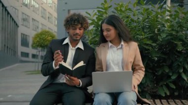 Hintli iş adamı ve Arap iş kadını iş ortağı şehir merkezindeki bankta oturan insanlar dizüstü bilgisayarla notlar yazıp konuşarak işlerini tamamlıyorlar.