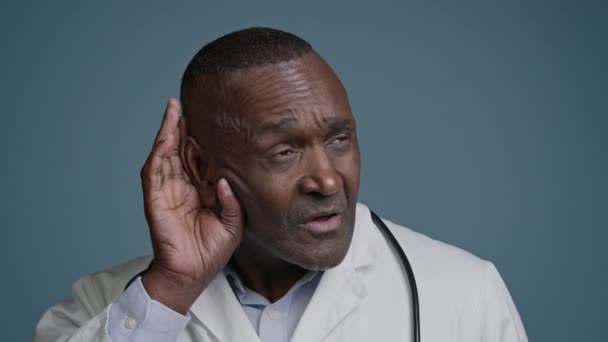 閉じ込められた混乱したアフリカ系アメリカ人男性男性医師スタジオブルーバックグラウンドで 耳の聴力損失問題の近くに手を保持して 静かなトーク広告医療テストを聞くために前方カメラを傾けます — ストック動画