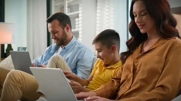 Addicted to modern technology dad mom son using different gadgets happy Caucasian family spend time together parents working remote on laptop at home on couch kid boy child playing video game on phone