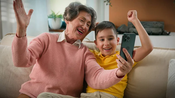 Caucasian grandmother and grandson together at home couch photographed selfie on mobile phone fool around funny family two generations grandma make vlog with kid boy taking pictures using smartphone