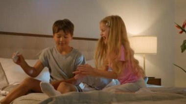 Two funny playful smiling Caucasian children boy girl playing together in bedroom bed evening brother teasing sister with flashlight kids siblings fooling around having fun happy family relationships